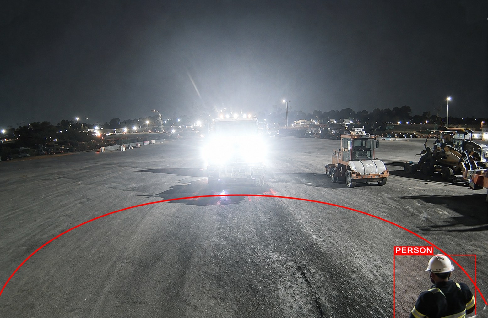 Night-time site camera detecting and boxing a worker in a hard hat at the edge of the danger zone, against the glare of an oncoming truck's headlights